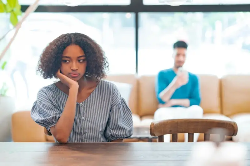 Une jeune femme songeuse, le menton appuyé sur la main, semble réfléchir à un **problème de couple à qui en parler**. Son compagnon est flou en arrière-plan.