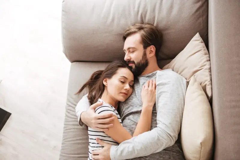 Un couple dort paisiblement sur un canapé. L'homme enlace la femme, une scène qui peut évoquer une baisse de libido chez l'homme, mais aussi un profond sentiment de sécurité et d'amour partagé.