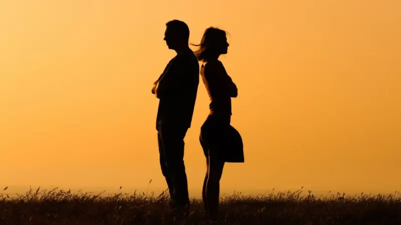Silhouettes d'un homme et d'une femme dos à dos, bras croisés, symbolisant la communication paradoxale dans un paysage au coucher du soleil.