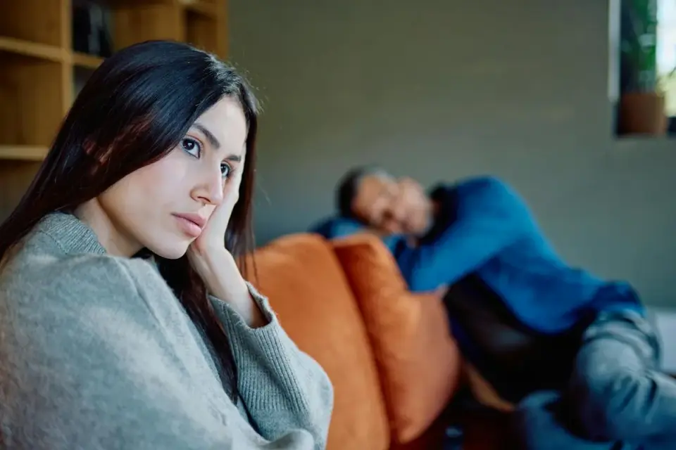 Une femme pensive, main sur la joue, face à un homme endormi. Le silence pèse, illustrant la psychologie du refus de communiquer dans le couple.